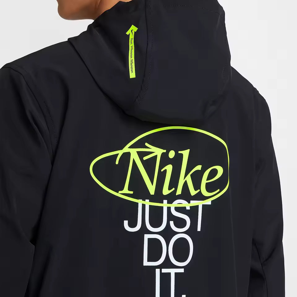 Close-up of the back of a black Nike hooded jacket with a large neon yellow-green outlined Nike logo and white 'JUST DO IT.' text, worn by a model against a light gray background.